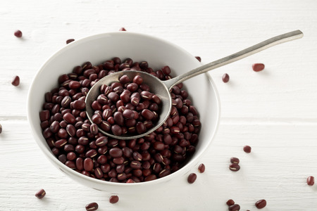 Raw, uncooked, dried adzuki (red mung) beans in white bowl with metal spoon on white wood table background with selective focusの写真素材