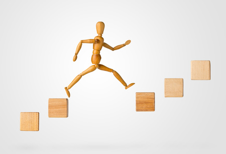 Wooden stick figure jumping from one wooden block on rising steps to the next - achievement, career or objective concept on white backgroundの写真素材