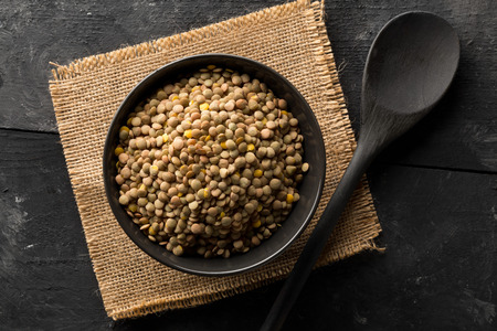 Raw, dry, uncooked brown lentil legumes in black bowl on rustic wood table background with selective focus top view flat lay from aboveの写真素材