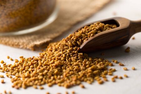 Dried, raw fenugreek seed in wooden scoop with storage glass jar on white wooden table background...
