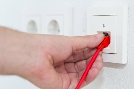 Man plugs red network cable in wall outlet for office or private home lan ethernet connection with power outlets flat view on white plaster wall background - selective focusの写真素材