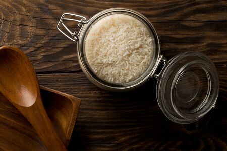 White uncooked, raw long grain rice in glass storage jar on rustic brown wooden table top view flat lay from above  - selective focusの写真素材