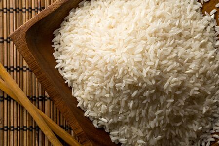 White uncooked, raw long grain rice in wooden bowl with chopsticks on bamboo mat top view flat lay from above - selective focusの写真素材