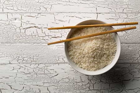 White uncooked, raw long grain rice in white bowl with chopsticks on rustic white wooden table with copy space top view flat lay from above - selective focusの写真素材