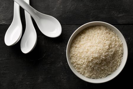 White uncooked, raw long grain rice in white bowl with spoons on black wooden table top view flat...