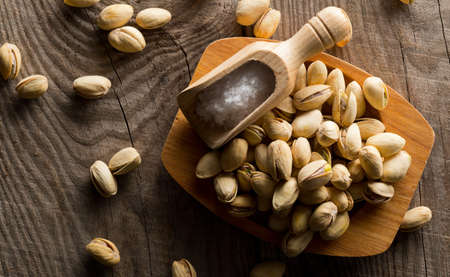 Heap of salted, roasted green pistachio nuts snack in wooden bowl on wood background with sea salt...