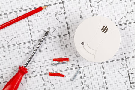 Smoke detector or fire alarm sensor on white architectural plans background with tools and screws, house safety or security concept, flat lay top view from aboveの写真素材
