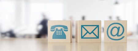 Wooden cube or box telephone, envelope letter and e-mail symbols in a row on wooden table with a blurred office in the background, contact us symbols or banner, 3D illustrationの写真素材