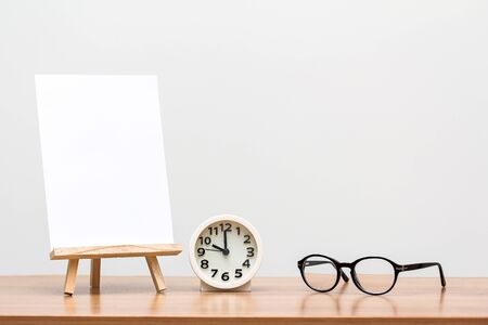 wooden easel with blank canvas, round clock and glasses on tableの写真素材