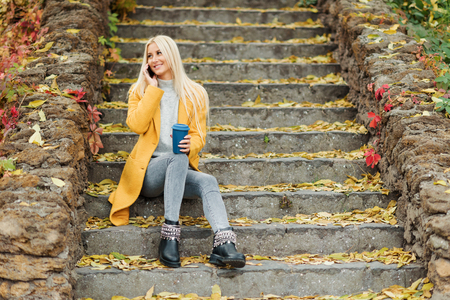 Young blond woman sitting in the city park and drinking coffee to go, using mobile phoneの写真素材