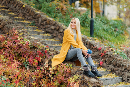 Stylish happy young blond woman in bright yellow coat holds a cup of coffee to go in the city park. Portrait of a young cheerful woman at work breakの写真素材