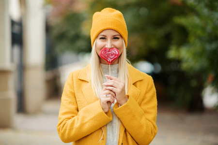 Saint Valentine day concept. Fashion portrait blond young woman in yellow coat having fun with red lollipop heart over street background.の写真素材