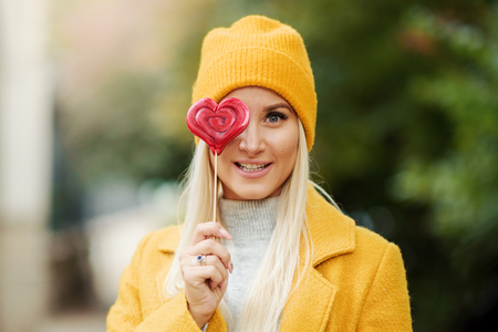 Saint Valentine day concept. Fashion portrait blond young woman in yellow coat having fun with red lollipop heart over street background.の写真素材