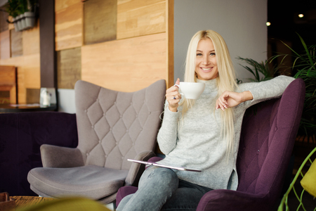 Reading Concept. Young blond Woman using Tablet and Drinking Coffee. Relaxing in Cozy cafe, Sit on the Chairの写真素材