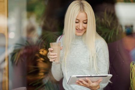 Reading Concept. Young blond Woman using Tablet and Drinking Coffee. Relaxing in Cozy cafe, Sit on the Chairの写真素材