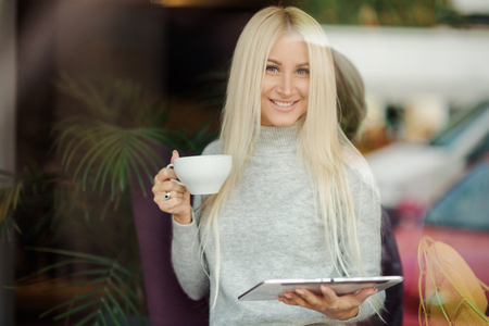 Reading Concept. Young blond Woman using Tablet and Drinking Coffee. Relaxing in Cozy cafe, Sit on the Chairの写真素材