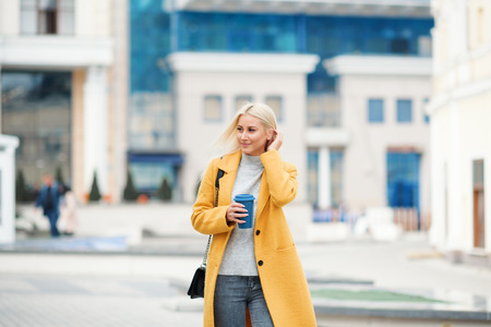 Young blond woman in a yellow bright coat with a cup of coffee to go, hurries to meet on a city streetの写真素材