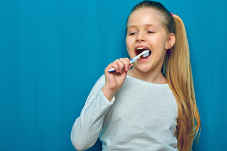 Little girl with long blonde hair cleaning teeth with toothy brush. Blue wall backgroundの写真素材