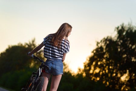 young girl posing standing with the bike on the road in the sunset timeの写真素材