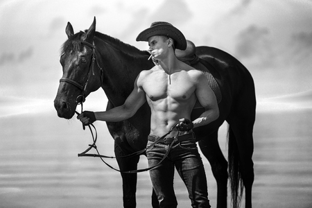 Macho man handsome cowboy with amazing muscles and abs and horse are on the background of sky and water. Western style. Black and white photoの写真素材