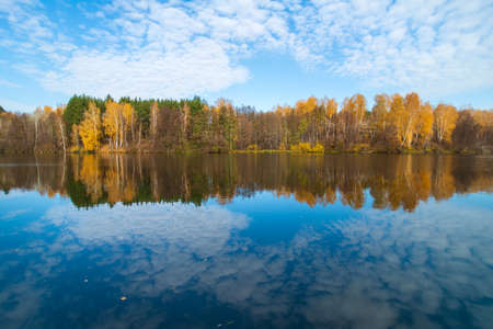 Beautiful autumn landscape with clear blue lake and yellow autumn trees. The sky with white clouds and autumn yellow trees are reflected in the mirror of the water.の写真素材