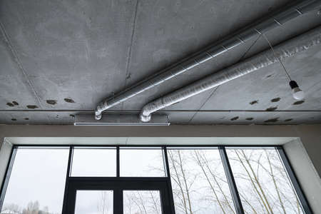 Wiring of ventilation pipes on a concrete ceiling in a house under construction. The room is under construction with a rough finish, plastered walls, concrete floor and ceiling.の写真素材
