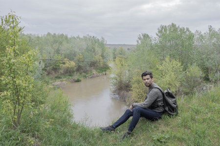 A man in nature, with a backpack sitting on the edge of a cliff, along the riverの写真素材