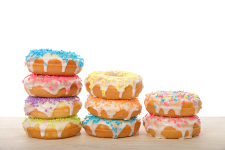 Many colorful frosted cake donuts with candy sprinkles stacked in rows laying on wood table isolated on white background. Fun festive party food.の写真素材