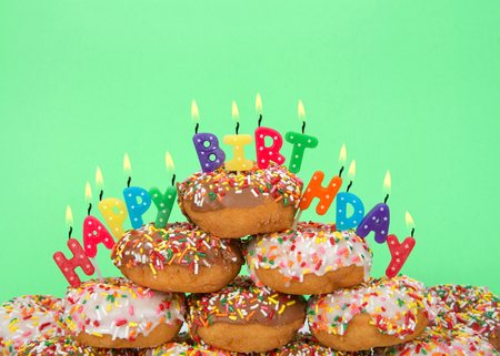 Chocolate and white frosted donuts covered in candy sprinkles piled into a cake pile with Happy Birthday candles burning brightly. Green background.の写真素材