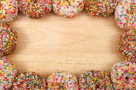 Flat lay view of many frosted donuts, chocolate and vanilla with candy sprinkles arranged in a border frame on a light wood table with copy space.の写真素材