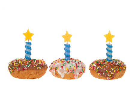 Vanilla cake donuts with white and chocolate frosting and colorful candy sprinkles lined up side by side, Blue decorative candles with stars on top isolated on white background.の写真素材