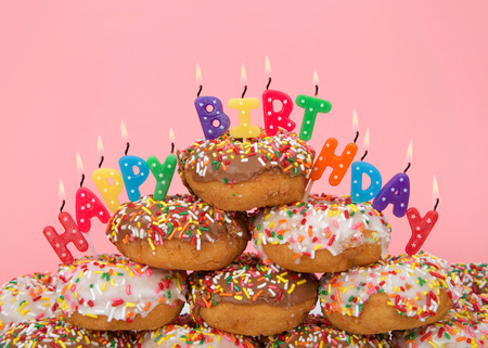 Chocolate and white frosted donuts covered in candy sprinkles piled into a cake pile with Happy Birthday candles burning brightly. Pink background.の写真素材
