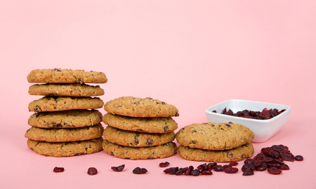 stacks of oatmeal cranberry cookies on a pink surface and background. Square bowl of dehydrated cranberries behind stacks with random cranberries on the tableの写真素材
