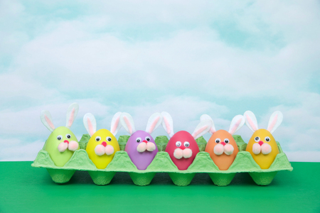 Colorful decorated Easter Eggs with bunny faces and ears sitting in an egg carton on green table, vintage sky background. Fun easter concept.の写真素材