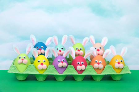 Colorful decorated Easter Eggs with bunny faces and ears sitting in an egg carton on green table, vintage sky background. Fun easter concept.の写真素材
