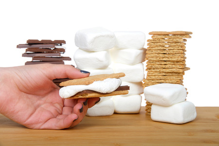 Young female hand holding homemade s'more with stacks of graham crackers, chocolate and marshmallows on a wood table, isolated background on white.の写真素材