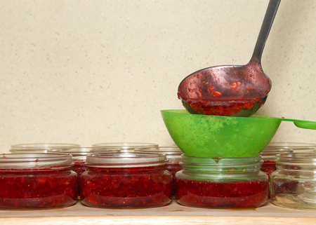 Close up on ladle pouring fresh hot homemade strawberry jam into glass jars through green plastic funnel. Wood table brown speckled wall background. Copy space.の写真素材