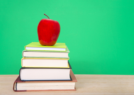 Hard bound books with various colored covers stacked on light wood table with red apple on top, dark green background similar to old fashioned chalk board. Back to school concept.の写真素材