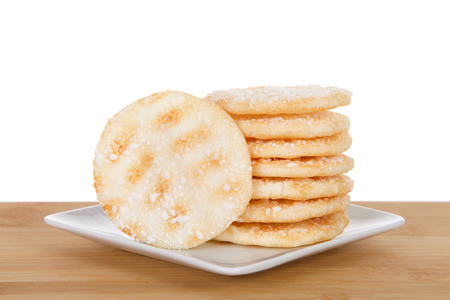 Pile of Japanese rice crackers on a small white square porcelain plate on wood table isolated behind. Rice crackers are usually low in sugar and fat.の写真素材