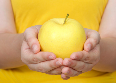 Young female hands holding one golden delicious yellow apple with yellow background.の写真素材