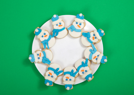 Snowman sugar cookies decorated with marshmallow fondant, blue royal icing scarf and hats arranged in a circle on a porcelain plate. Green background.の写真素材