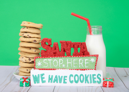 Chocolate chip cookies stacked 12 high on a small round plate, Glass bottle of milk with straw next to sign SANTA STOP HERE WE HAVE COOKIES. Light wood table green background. light wood tableの写真素材
