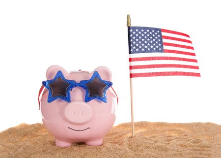 Traditional pink piggy bank wearing star glasses sitting in sand with American Flag waving, isolated on white.の写真素材