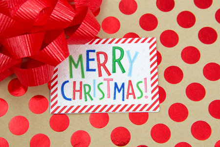 Top view flat lay close up of a colorful Merry Christmas label on a red polka dot on brown paper covered present with a red bow.の写真素材