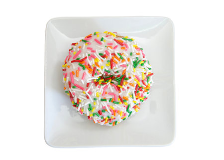 Top view flat lay of one vanilla cake donut with vanilla frosting covered in bright colorful candy sprinkles on a small square off white porcelain plate, isolated on whiteの写真素材
