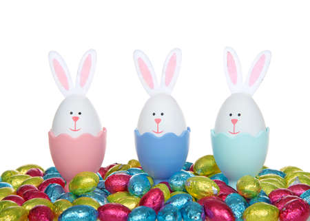 Row of Easter Eggs crafted into bunnies with felt ears, sitting in colorful egg cups with colorful foil wrapped chocolate egg candies, isolated on white background.の写真素材