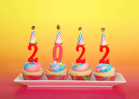 Vanilla cupcakes frosted with pink and blue buttercream frosting on a rectangular plate pink background and yellow with pink 2022 candles wearing party hats. Happy New Year theme.の写真素材
