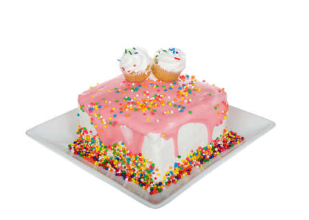 White vanilla frosted square cake on a square porcelain plate, dripping with strawberry ganache and rainbow sprinkles. Two cupcakes side by side on top. Isolated on white.の写真素材