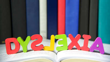 Bright colorful wooden toy blocks on an open book spelling dyslexia. Learning challenges disabilities. S, L and E are backwards. Hard bound books lined up in backgroundの写真素材