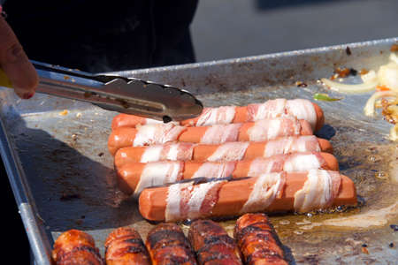 Street vendor cooking bacon wrapped hot dogs, tongs turning food. Popular cuisine for street fairs and eventsの写真素材
